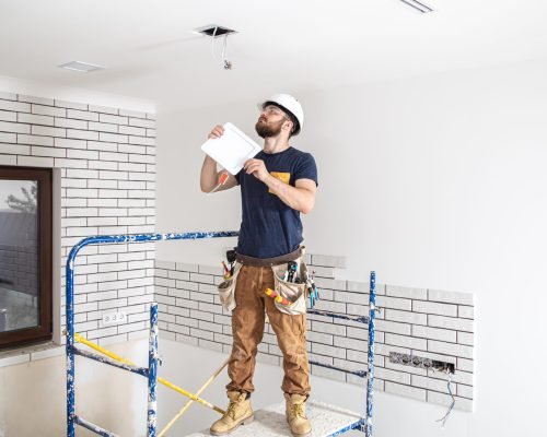 Electrician Builder with beard worker in a white helmet at work, installation of lamps at height. Professional in overalls with a drill on the background of the repair site.